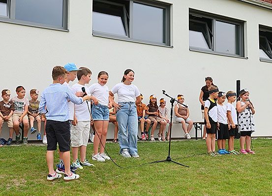 Eine Gruppe von Kindern und Erwachsenen auf einem Rasenfeld, einige stehend und andere auf einer Bank sitzend, wahrscheinlich bei einer Talentshow mit Mikrofonen.