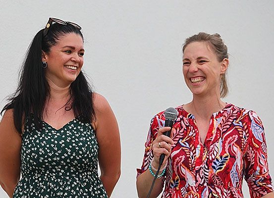 Zwei Frauen stehen draußen, eine hält ein Mikrofon und beide lächeln. Die Frau auf der linken Seite trägt ein grünes Kleid mit weißen Blumen. Die Frau auf der rechten Seite trägt ein rotes Blumenhemd.
