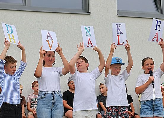 Fünf Kinder halten Buchstaben D, H, A, V, N, A, K, L, E, A vor einem Gebäude hoch, während eine Menge zuschaut.