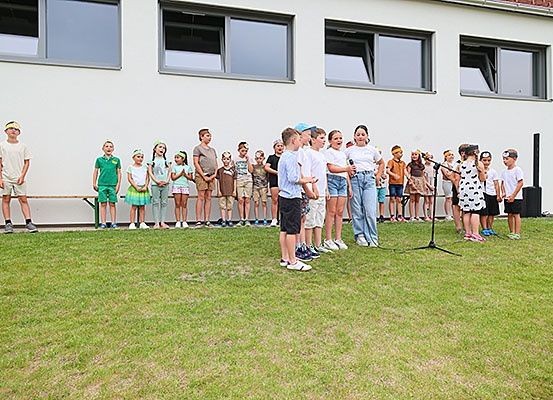 Kinder stehen in zwei Reihen vor einem Gebäude mit Fenstern und singen. Ein Mikrofon auf einem Ständer steht vor ihnen.