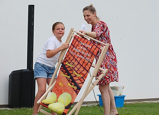Eine Frau und ein Mädchen halten einen hölzernen Stuhl mit Tennisthema in einem Garten. Die Frau lächelt und schaut das Mädchen an. Ein Tennisschläger und Bälle sind auf dem Stuhl.