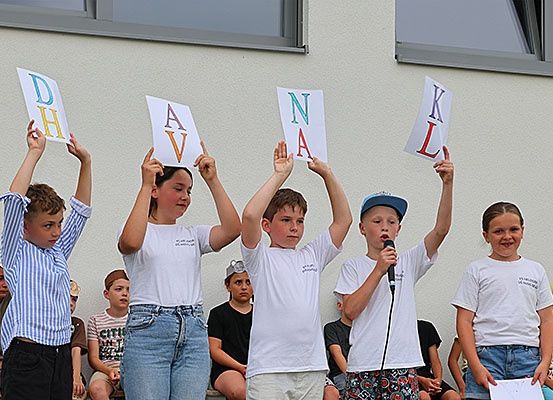 Fünf Kinder, die die Buchstaben 'D', 'A', 'V', 'N', 'A' und 'K' hochhalten, stehen vor einem Gebäude, mit einem Jungen, der ein Mikrofon hält. Dahinter sitzen mehrere Personen auf einer Bank und beobachten.