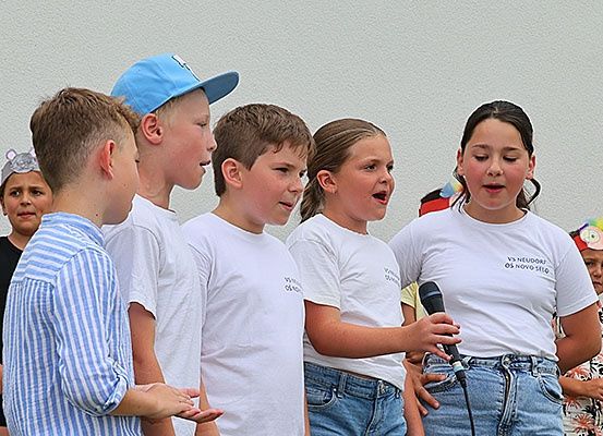 Eine Gruppe von Kindern, alle in weißen T-Shirts gekleidet, singen ein Lied mit einem Mikrofon. Ein Mädchen mit lockigem Haar singt, während ein anderes Mädchen das Mikrofon hält. Hinter ihnen steht eine Wand.