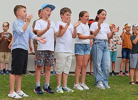 Eine Gruppe von Kindern in verschiedenen Kostümen steht auf einem Rasen und singt, einer hält ein Mikrofon.