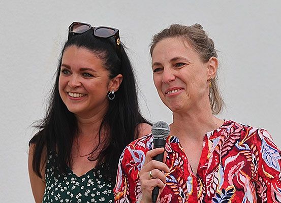 Zwei Frauen stehen nebeneinander, lächeln und eine hält ein Mikrofon. Die Frau auf der linken Seite hat eine Sonnenbrille auf dem Kopf und eine Halskette. Die Frau auf der rechten Seite trägt einen Ring und ein Blumenhemd. Sie scheinen sich bei einer Veranstaltung im Freien zu befinden.
