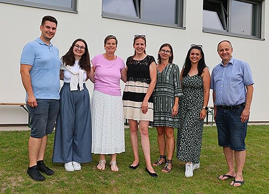 Eine Gruppe von sieben Personen, darunter zwei Männer und fünf Frauen, posiert für ein Foto vor einem weißen Gebäude mit Glasfenstern. Sie stehen auf einem Rasen und alle lächeln.