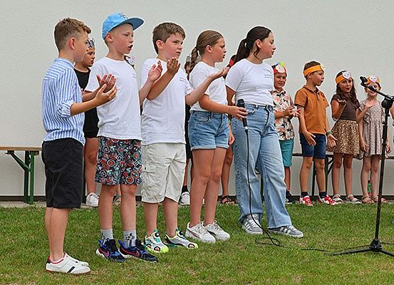 Eine Gruppe von Kindern singt auf einem Grasfeld mit einem Mikrofon vor ihnen. Einige von ihnen tragen Stirnbänder.