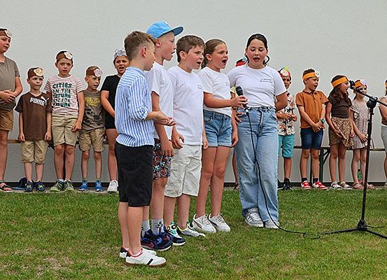 Eine Gruppe von Kindern in weißen T-Shirts mit Stirnbändern steht auf einer Bühne mit einem Mikrofon vor ihnen.