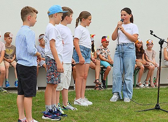 Eine Gruppe von Kindern mit Hüten und Sneakers steht auf einem Rasenfeld. Ein Mädchen hält ein Mikrofon in der Hand, wahrscheinlich singt sie.