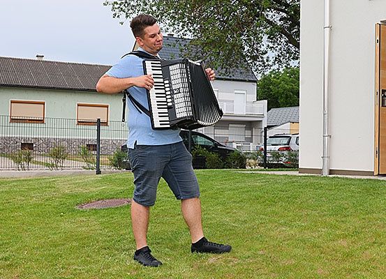 Ein Mann spielt ein Akkordeon auf einem Rasen vor einem Haus.