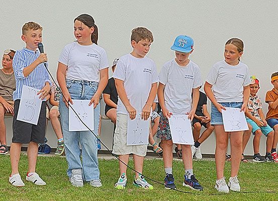 Eine Gruppe von Kindern steht auf einem Rasenfeld, hält Papiere und trägt Mützen und Turnschuhe. Ein Kind hält ein Mikrofon und spricht.