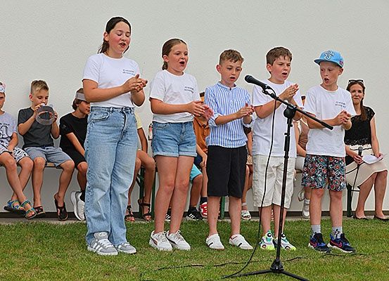 Eine Gruppe von Kindern tritt auf einer Bühne mit einem Mikrofon auf, während einige auf Bänken hinter ihnen sitzen und eine Frau ein Papier hält.