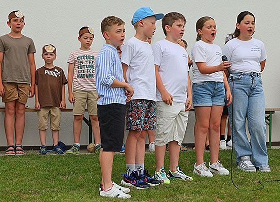 Eine Gruppe von Kindern mit Stirnbändern steht in einer Reihe und singt auf einem Rasenfeld, wobei zwei Mädchen ein Mikrofon halten. Dahinter sind zwei Holzbänke aufgestellt.