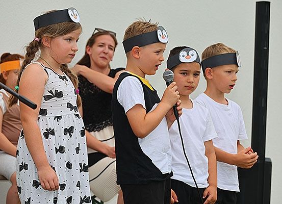 Eine Gruppe von Kindern mit Pinguin-Kopfbändern führt eine Aufführung durch, ein Junge hält ein Mikrofon. Hinter ihnen beobachtet eine erwachsene Frau, und ein Mädchen in einem schleifenbesetzten Kleid steht links.