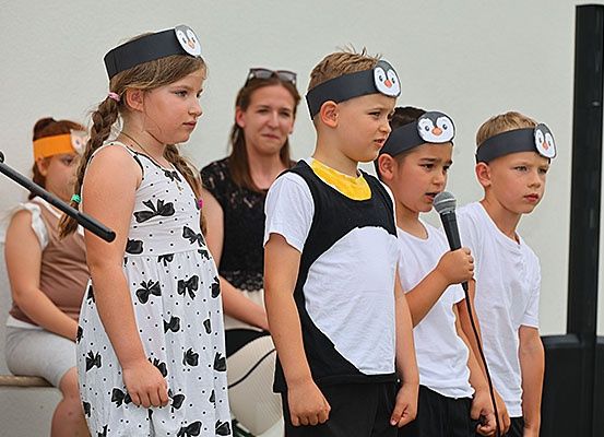 Eine Gruppe von Kindern mit Pinguin-Kopfbändern steht vor einem Mikrofon und führt wahrscheinlich eine Aufführung durch. Hinter ihnen beobachtet eine erwachsene Frau. Die Umgebung scheint eine Bühne mit einer weißen Wand und einer schwarzen Säule zu sein.