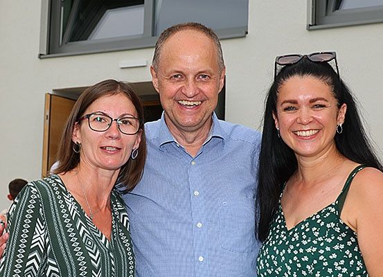Drei Personen, zwei Frauen und ein Mann, stehen eng beieinander und lächeln für ein Foto vor einem Gebäude.