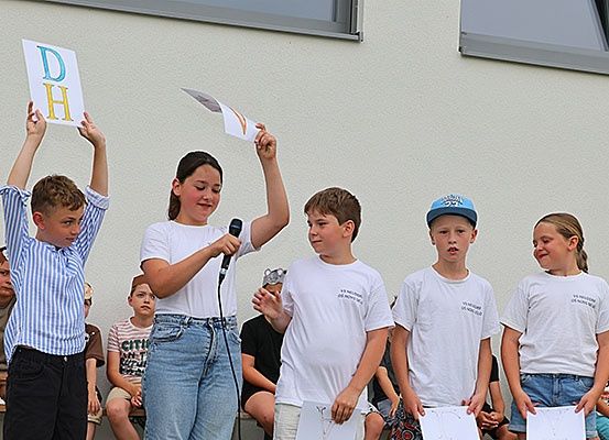 Fünf Kinder, die Papier und ein Mikrofon halten, stehen vor einem Gebäude. Sie führen möglicherweise eine Aufführung durch. Das Mädchen in der Mitte hält ein Mikrofon und Papier. Hinter ihnen sitzen Menschen auf einer Bank.