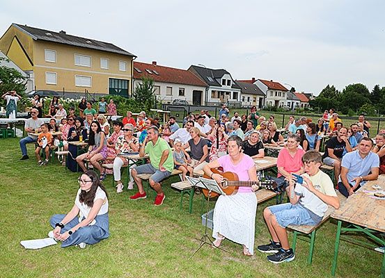 Eine Menschenmenge versammelt sich auf einer grasigen Freifläche und beobachtet eine Frau, die Gitarre spielt. Einige sitzen, andere stehen. Dahinter stehen Häuser in einer Reihe.