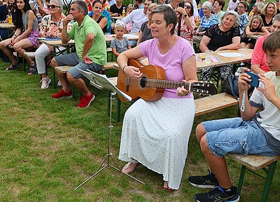 Eine Frau spielt Gitarre auf einer Bühne vor einem Publikum, während ein Junge vor ihr mit seinem Handy aufnimmt. Das Publikum sitzt auf Bänken.
