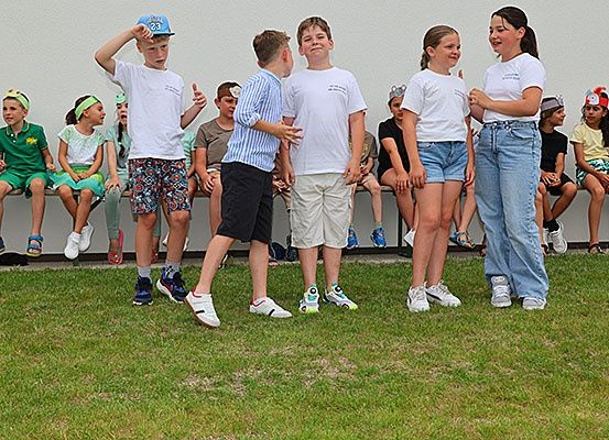Eine Gruppe von Kindern in Freizeitkleidung steht auf einem Außengelände, möglicherweise bei einer Schulveranstaltung. Einige von ihnen tragen Stirnbänder. Hinter ihnen sitzen einige Kinder auf einer Bank. Der Boden ist mit Gras bedeckt.