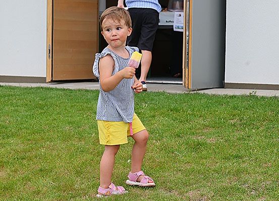 Ein kleines Mädchen steht auf dem Rasen, hält einen Eislutscher und schaut nach vorne. Hinter ihr steht eine Person in der Nähe der offenen Tür.