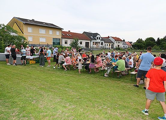 Eine große Gruppe von Menschen versammelt sich im Freien, einige sitzen auf Bänken, andere stehen. Die Umgebung ist ein Park mit grünem Rasen und mehreren Häusern im Hintergrund.