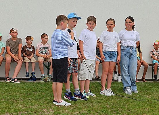 Eine Gruppe von Kindern steht auf einem grünen Rasen. Einige sitzen auf einer Bank, während andere stehen. Sie tragen alle Freizeitkleidung. Ein Kind hält ein Mikrofon und scheint zu sprechen.