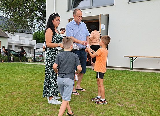 Eine vierköpfige Familie steht vor einem Gebäude und erhält Gegenstände von einem Mann in formeller Kleidung. Dahinter steht ein Gebäude mit offener Tür und einer Bank. Im Hintergrund Häuser und geparkte Autos.
