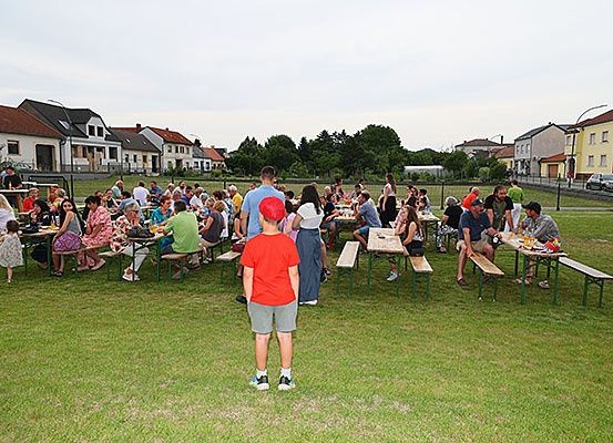 Eine Gruppe von Menschen versammelt sich auf einem Rasenfeld für ein Picknick, sitzt um Holztische mit Bänken herum, unter einem bewölkten Himmel.