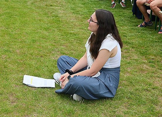 Eine Frau mit Brille sitzt auf dem Rasen und schaut nach oben. Sie trägt eine Uhr am Handgelenk und ein Papier liegt neben ihr. Zwei Personen sitzen hinter ihr.