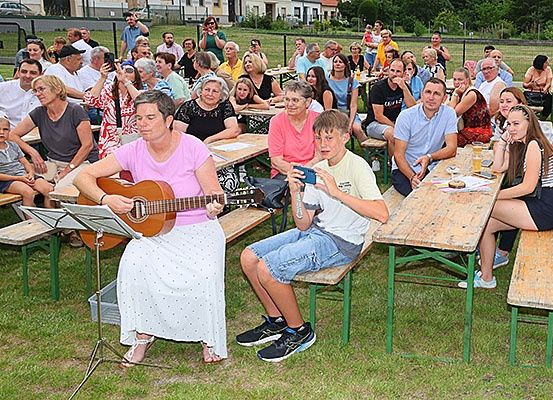 Eine Gruppe von Menschen sitzt um Picknicktische in einem Park herum. Einige genießen Getränke, während andere einer Frau beim Gitarrespielen zuschauen. Es sind Kinder anwesend, und die Atmosphäre ist entspannt und fröhlich.