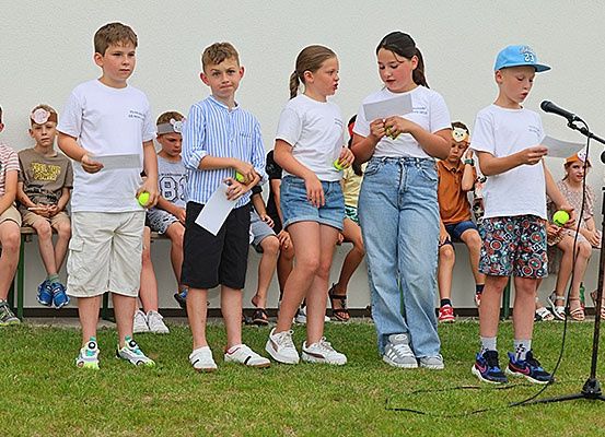 Mehrere Kinder halten Tennisbälle und Papiere in der Hand und stehen vor einem Mikrofon auf einem Rasenfeld. Dahinter sitzen mehrere Kinder auf einer Bank. Die Kinder vor ihnen führen wahrscheinlich auf. Die Wand hinter ihnen hat eine weiße Farbe.
