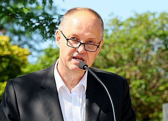 Ein Mann mit Brille und schwarzem Anzug steht an einem Podium und spricht in ein Mikrofon. Bäume und Pflanzen sind im Hintergrund.
