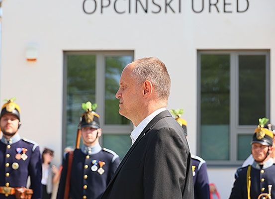 Ein Mann in einem Anzug steht vor einem Gebäude namens Opcinski Ured mit uniformierten Personen hinter ihm. Die Uniformen haben dekorative Federn und Medaillen. Das Gebäude hat Fenster und eine Lampe.