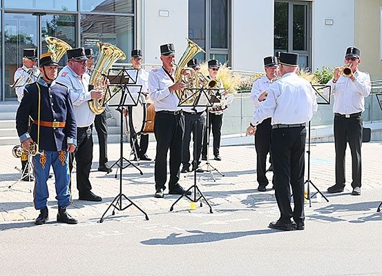 Eine Blaskapelle in traditionellen Uniformen steht auf einem gepflasterten Bereich und spielt Instrumente, mit einem Gebäude und Grünpflanzen im Hintergrund.