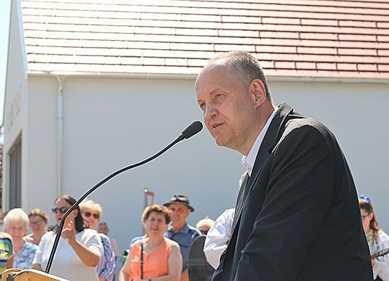 Ein Mann in einem Anzug spricht an einem Rednerpult mit einem Mikrofon vor einer Menschenmenge, von der einige Hüte und Brillen tragen. Im Hintergrund befindet sich ein Gebäude mit einem roten Dach.