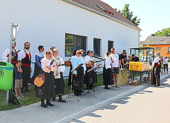 Eine Gruppe von Menschen in traditionellen Kostümen spielt Saiteninstrumente auf einem Bürgersteig. Hinter ihnen steht ein Gebäude, ein Mülleimer und eine kleine Menge Menschen.
