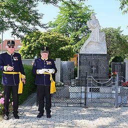 Zwei Männer in Militäruniformen stehen neben einer Gedenkstatue in einem Park. Das Denkmal ist von einem Zaun umgeben.