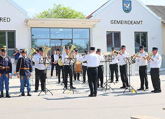 Eine Blaskapelle spielt vor einem Gebäude, wahrscheinlich einem Rathaus, mit einem Logo an der Fassade. Die Musiker sind in Uniformen und Hüten gekleidet.