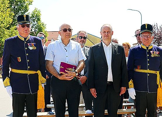 Eine Gruppe von Männern in Uniformen und Anzügen, einschließlich Medaillen, stehen im Freien mit einer Bank und Bäumen im Hintergrund.