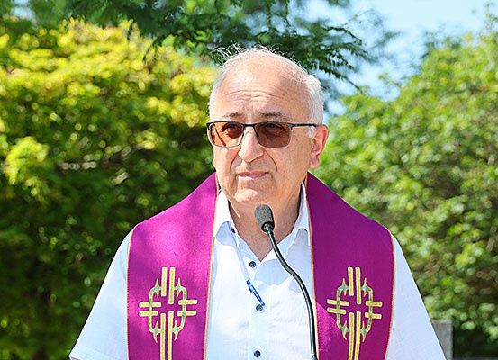 Ein älterer Mann in einem weißen Hemd und einem violetten Stola steht an einem Rednerpult mit einem Mikrofon vor ihm. Er trägt eine Sonnenbrille und scheint eine Rede zu halten. Dahinter befinden sich verschwommene Bäume und ein blauer Himmel.