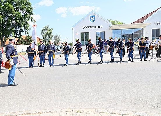 Eine zeremonielle Militärparade findet vor einem Gebäude mit dem Schild Opčinski Ured statt, mit Musikern, die spielen, und einer zuschauenden Menge.