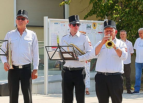 Drei Männer in weißen Uniformen mit schwarzen Mützen spielen Trompeten auf der Straße, mit einem Notenständer und Noten vor ihnen. Dahinter stehen Menschen, einer mit Brille und einer mit einer Uhr. Bäume und ein Gebäude sind im Hintergrund.