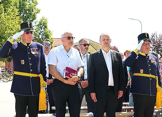 Eine Gruppe von Menschen steht draußen auf Bänken, zwei in Militäruniformen salutieren. Der Mann im weißen Hemd hält eine lila Mappe.
