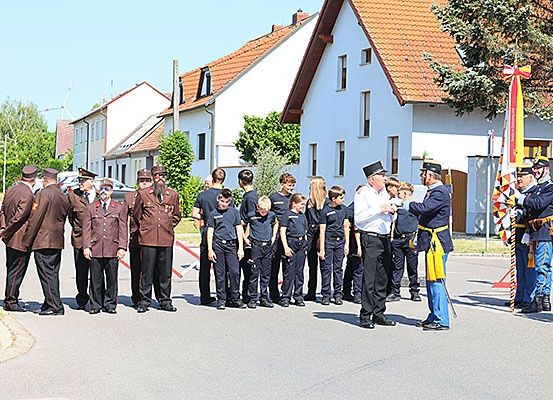 Eine Gruppe uniformierter Personen steht in einer Reihe, möglicherweise eine feierliche Veranstaltung. Sie stehen vor einem Gebäude mit roten Dächern. Die Männer vorne tragen Hüte.