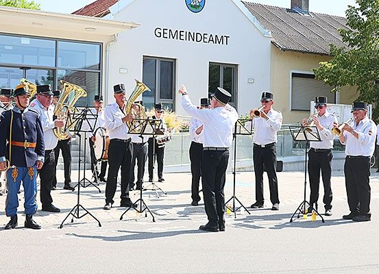 Eine Gruppe Musiker, einige in Uniform, spielen Instrumente vor einem Gebäude mit der Aufschrift 'Gemeindeamt'.