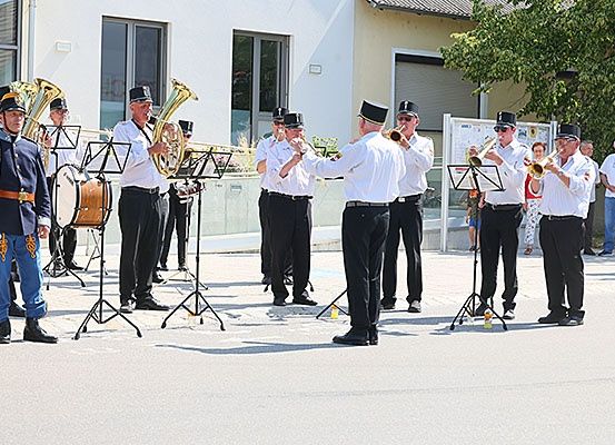 Eine Band spielt im Freien, wobei Musiker Blechblasinstrumente und Trommeln spielen, während einige Zuschauer zusehen.