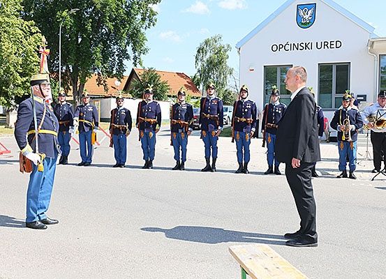 Eine Militäreinheit in blauen Uniformen steht in einer Reihe vor einem Gebäude mit dem Schild 'Opcinski Ured'.