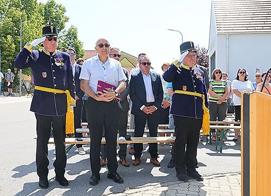 Zwei Männer in Uniform salutieren vor einer Menge. Ein Mann in einem weißen Hemd hält eine violette Mappe. Ein Rednerpult steht in der Nähe.