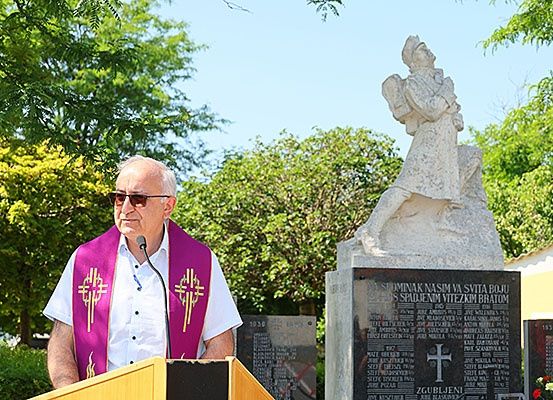 Ein älterer Mann in priesterlicher Kleidung steht an einem Podium und spricht in ein Mikrofon, mit einer Statue hinter ihm. Die Statue stellt eine Person mit Flügeln dar, und die Umgebung ist mit grünen Bäumen und Büschen geschmückt.
